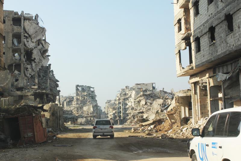 On 24 January 2015 in the Syrian Arab Republic, vehicles of the United Nations travel a dusty road lined with the rubble remnants of destroyed buildings, in the Old City of Homs.